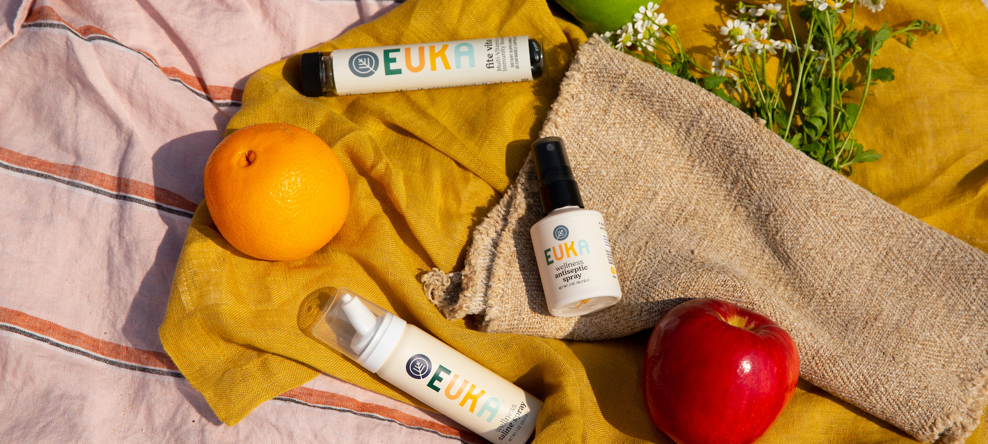 Euka Wellness Antiseptic Spray, Wellness Saline Spray, and Fite Vite laying on a picnic blanket next to an apple